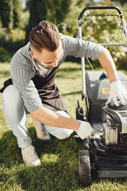 A man wearing safety glasses and a checkered shirt is kneeling on a well-maintained grassy lawn, inspecting or performing maintenance on a lawn mower. The garden features lush green grass, with neatly